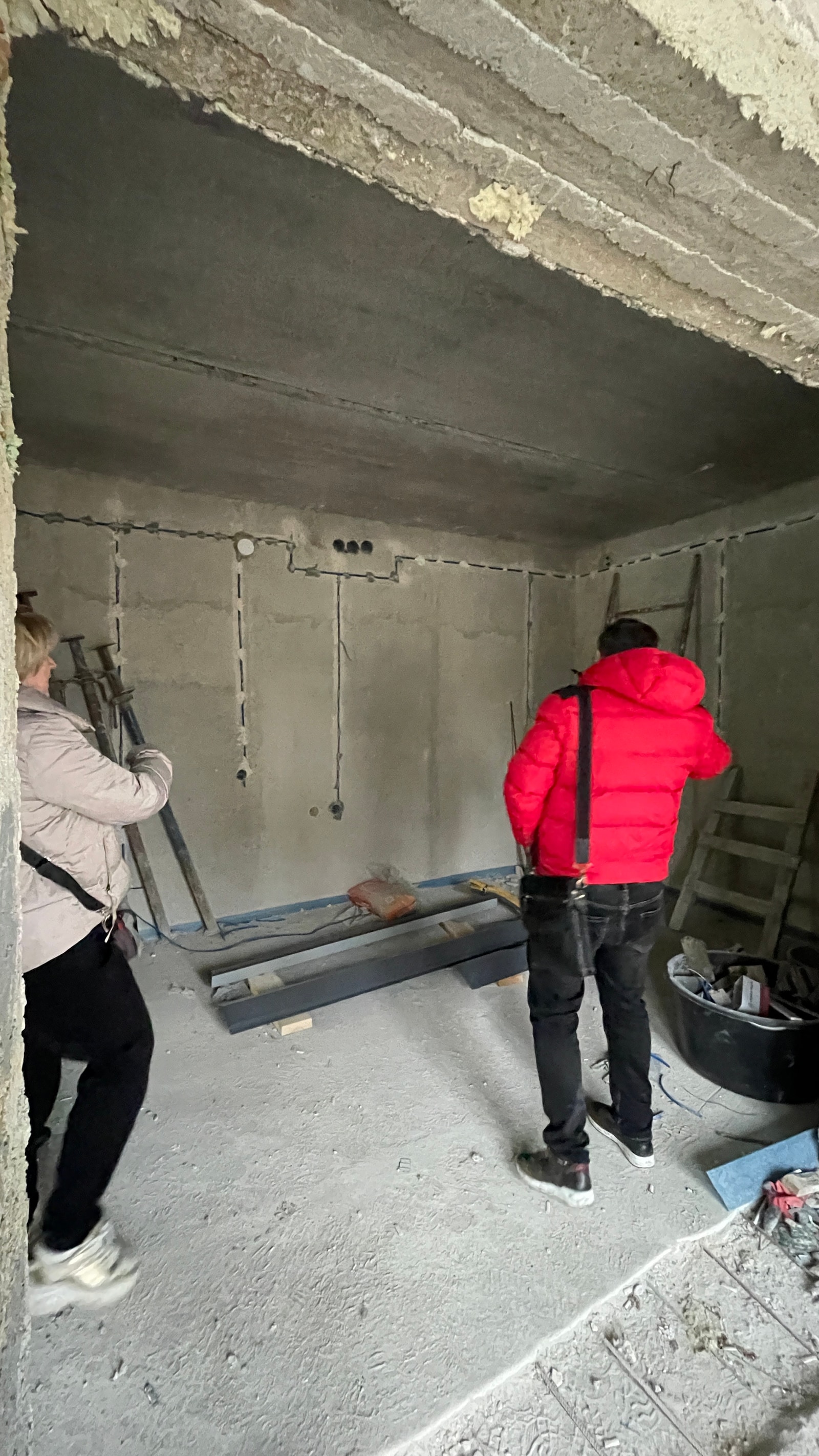 Двое — женщина и мужчина в красной куртке — осматривают голые бетонные стены помещения под кухню