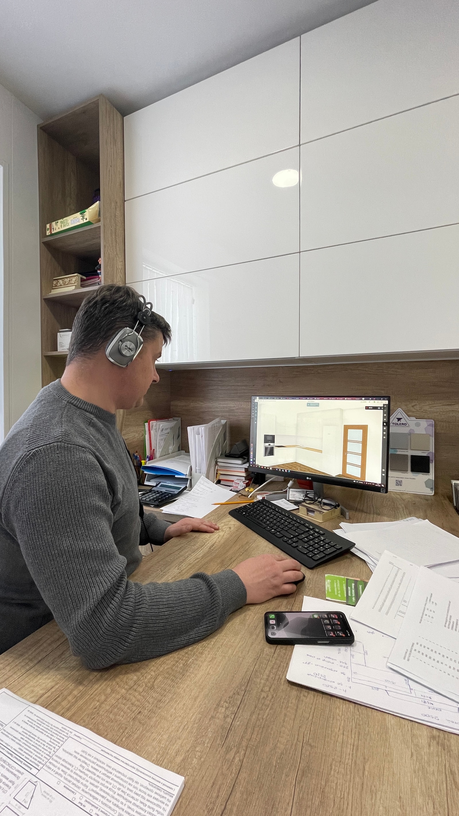 Сотрудник за компьютером проектирует кухню в 3D-программе, рабочий стол с чертежами и документами