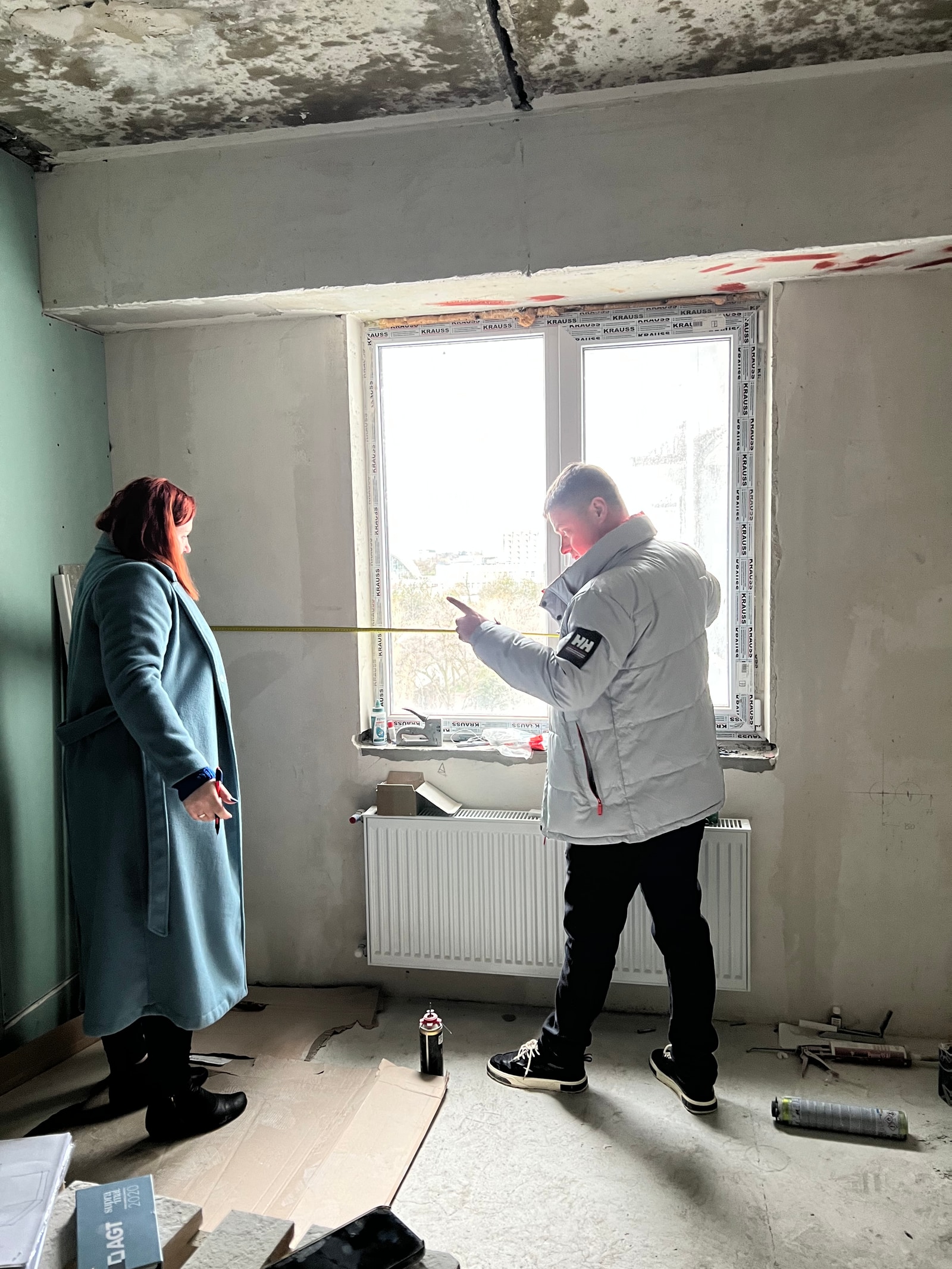 Замер у окна: мужчина с рулеткой объясняет женщине-клиенту планировку в пустой черновой квартире