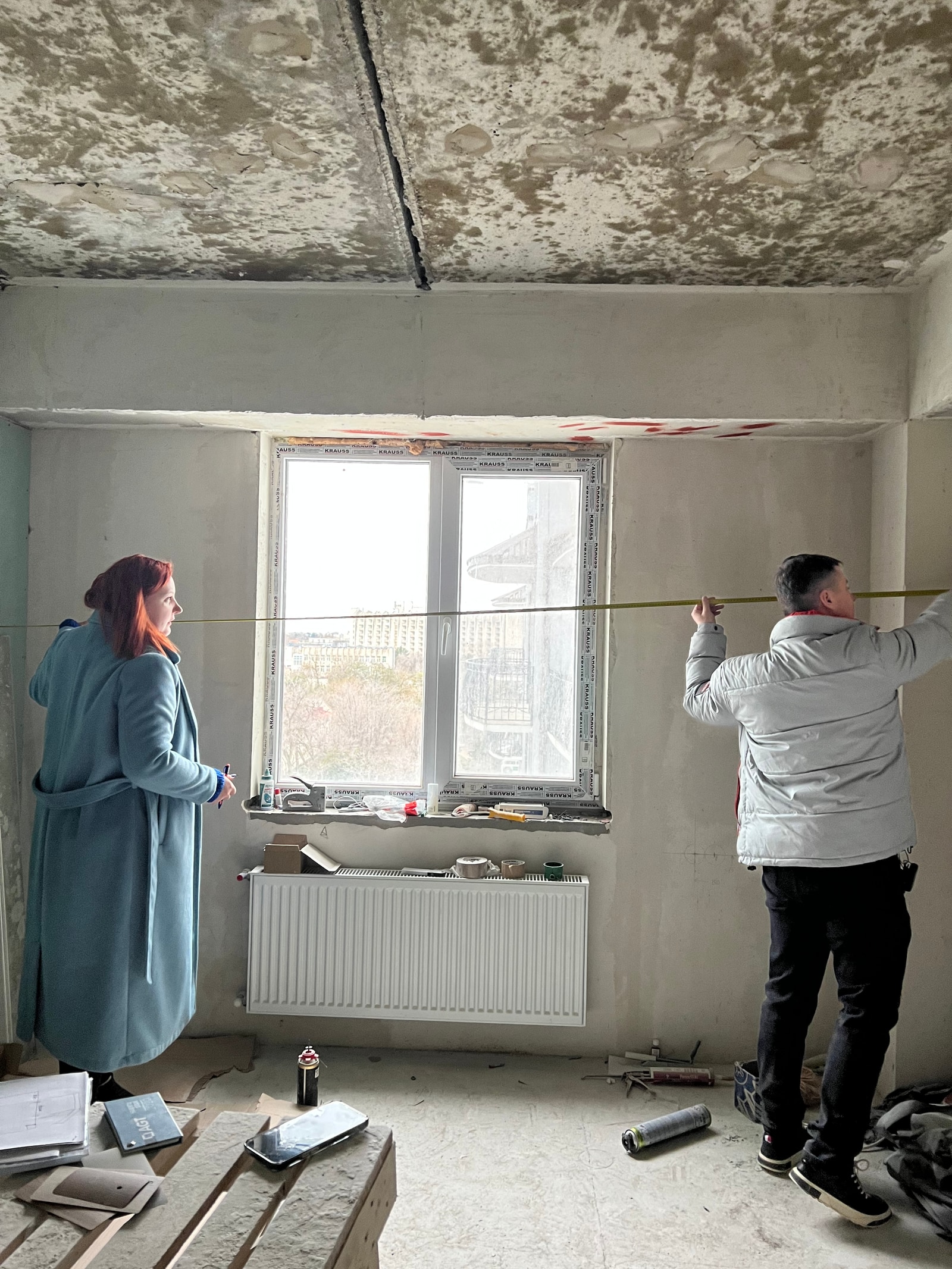 Замерщик с рулеткой и женщина-клиент в пустой квартире под ремонт, голые стены и потолок