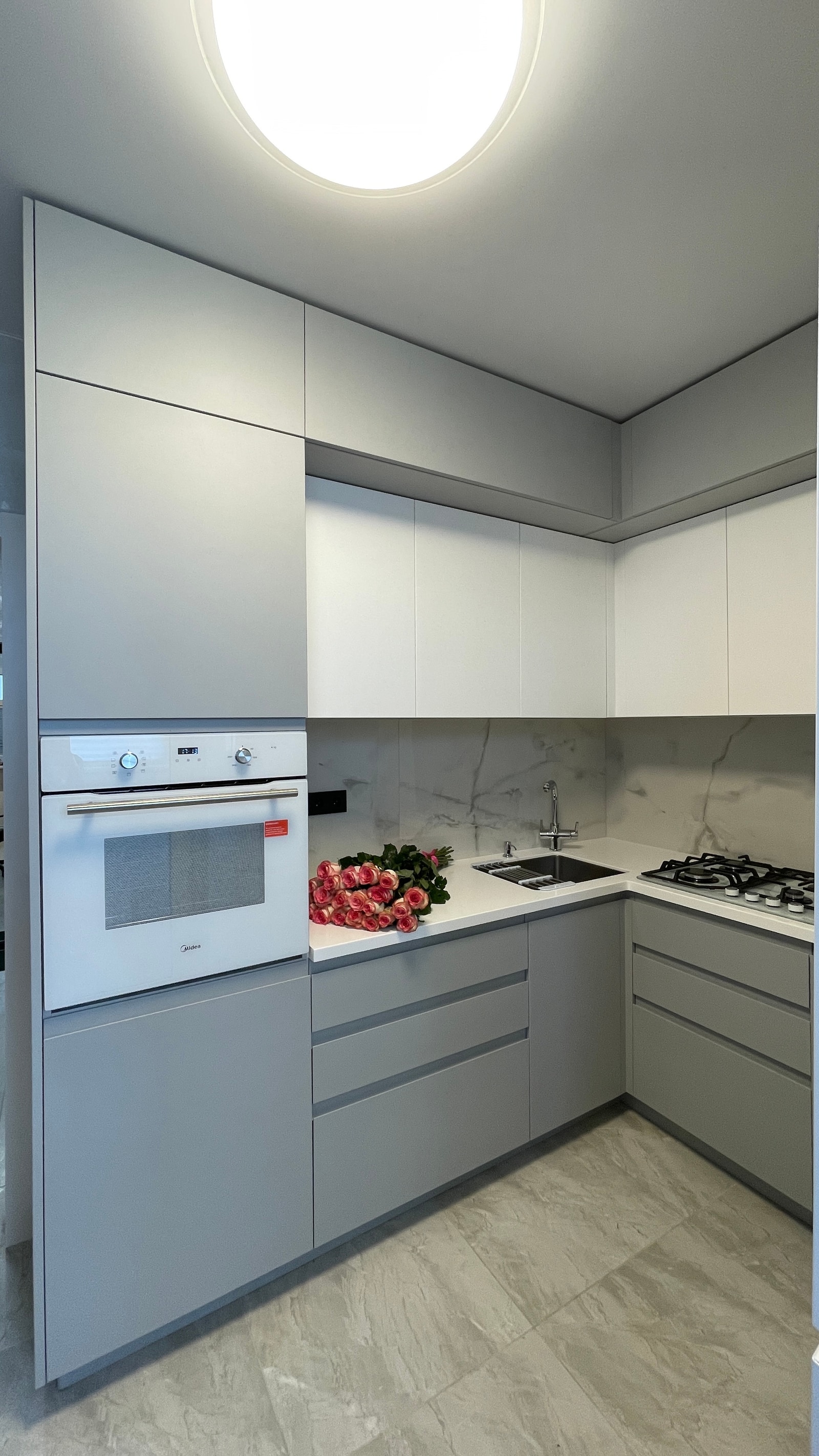 Угловая серо-белая кухня с белой духовкой в пенале, газовой плитой и мраморным фартуком, общий план с потолочным светильником