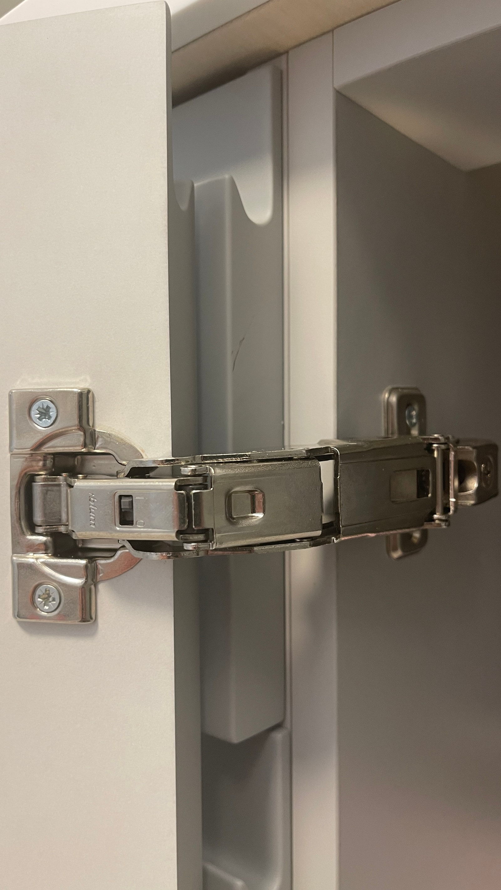 Крупный план хромированной петли Blum на белом матовом фасаде, видна внутренность шкафа