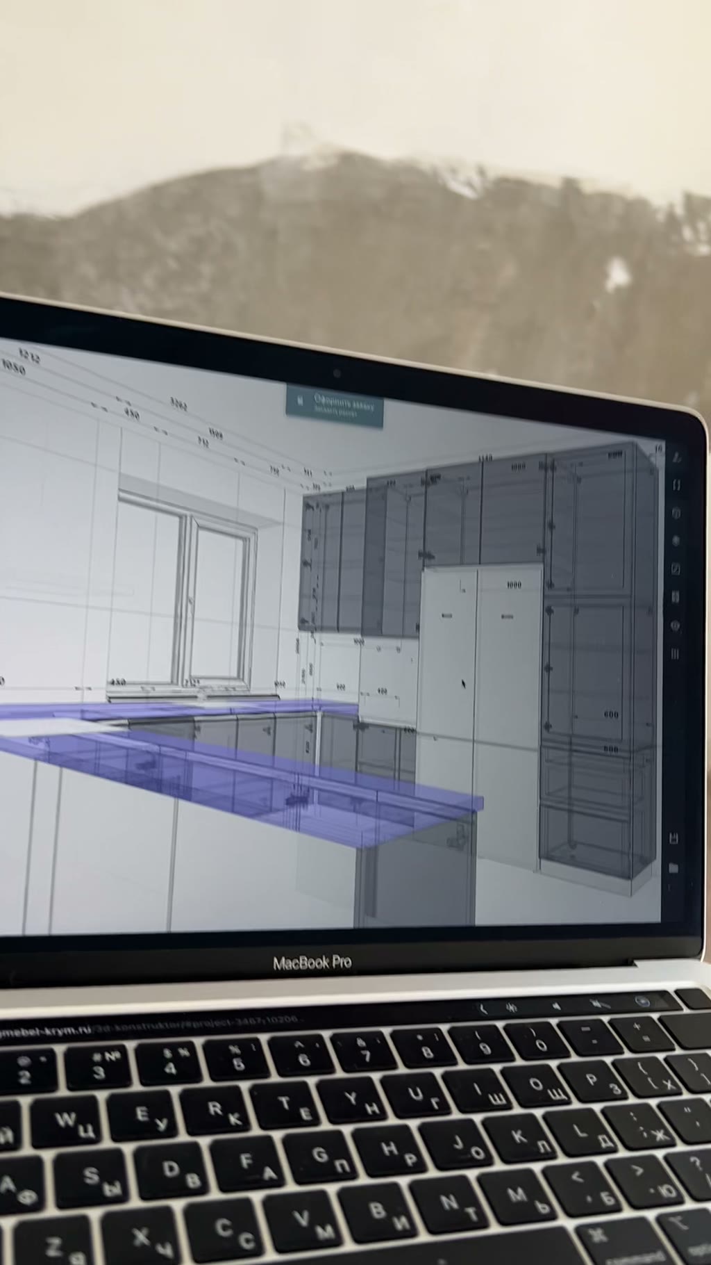 MacBook Pro с открытым 3D-проектом кухни в конструкторе: верхние и нижние шкафы, выделенная синим барная стойка, размерные линии, окно на фоне стены в ремонте