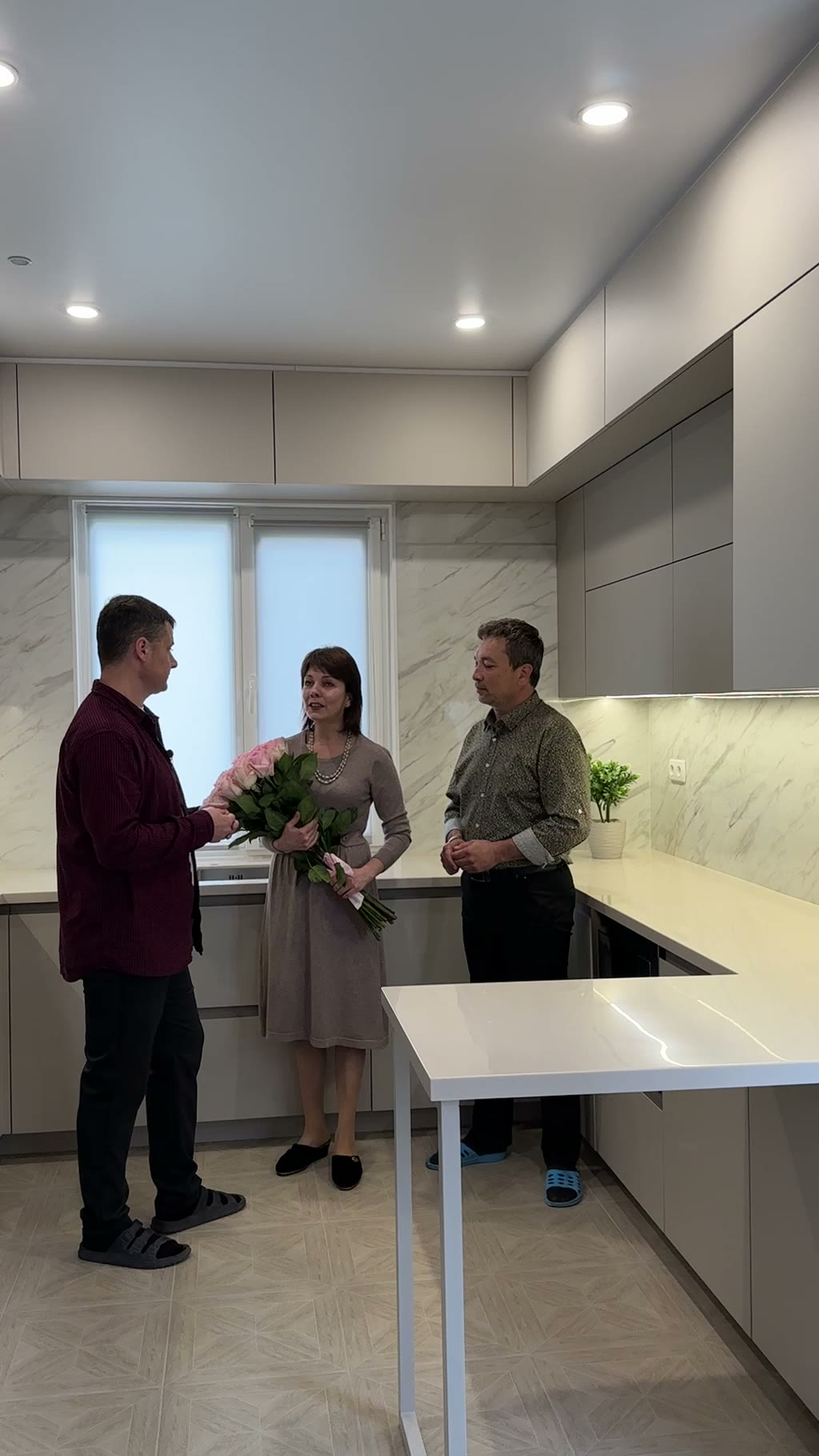Трое клиентов на сдаче готовой угловой кухни: женщина с букетом розовых роз, мраморный фартук, серые матовые фасады без ручек, белая столешница-стол