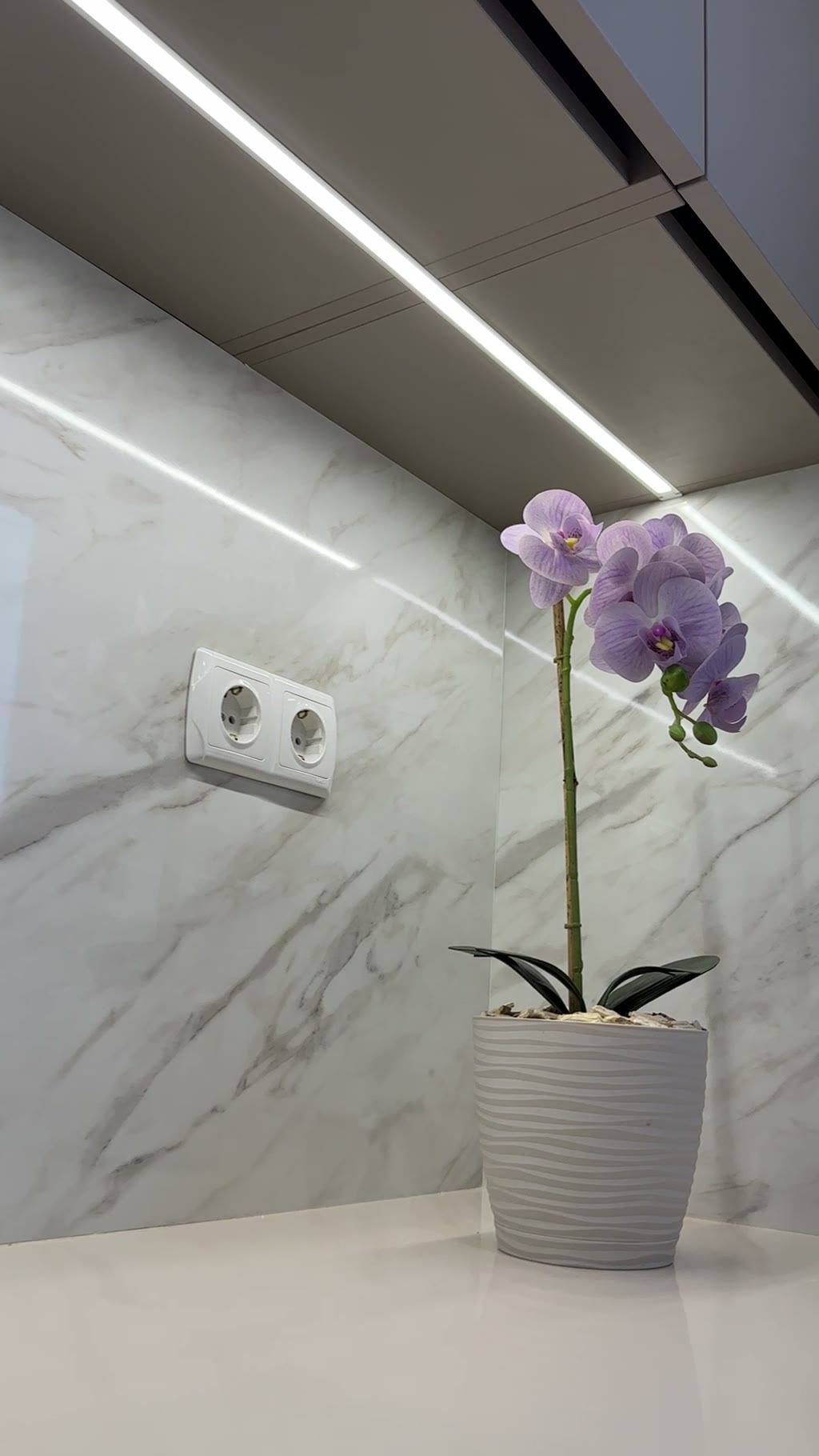Кухонный фартук под мрамор с LED-подсветкой снизу верхних шкафов, на белой столешнице горшок с сиреневой орхидеей, двойная розетка на стене
