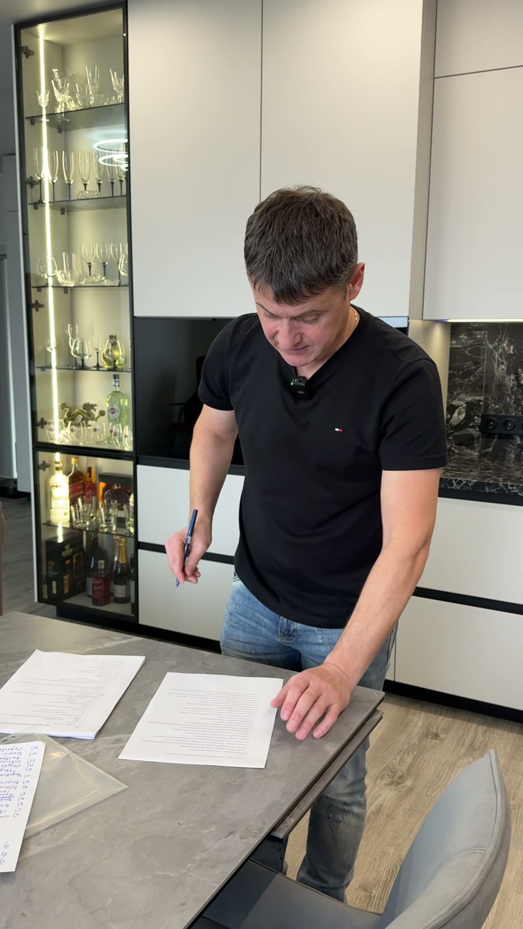 Мужчина в чёрной футболке подписывает договор на столешнице; фон — белая кухня с витриной-баром, хрусталём и бутылками алкоголя, тёмный мраморный фартук