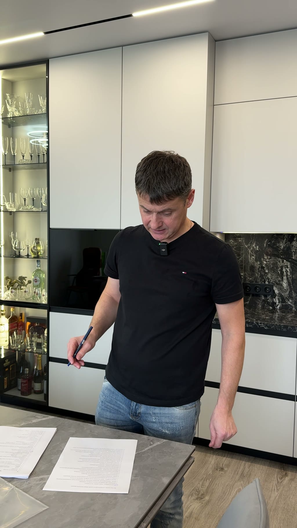 Мужчина с ручкой у столешницы подписывает договор на фоне белой кухни с витриной, бокалами и мраморным фартуком