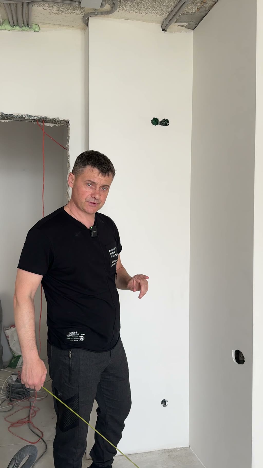 Замерщик с рулеткой на объекте в стадии ремонта: белые стены, открытая проводка, подрозетники, красный кабель на полу