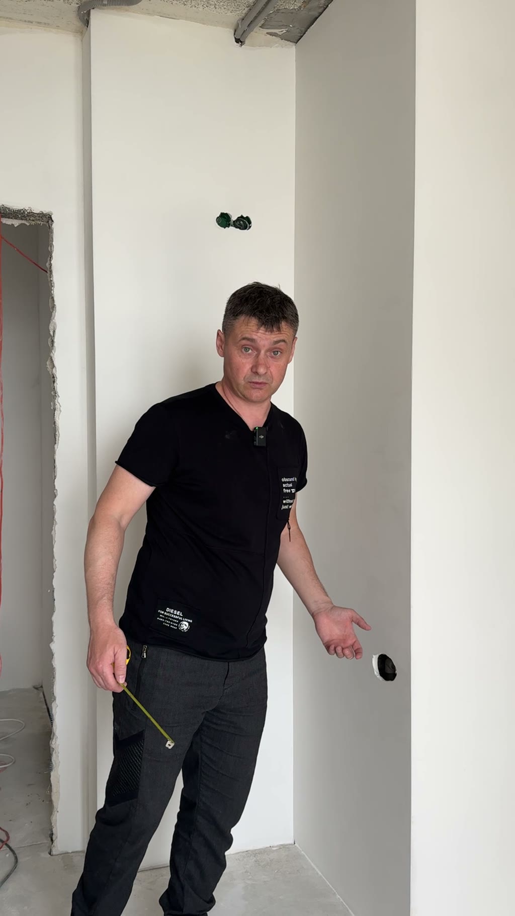Замерщик в чёрной футболке с рулеткой в руках на объекте в стадии ремонта: белые стены, розетки, красная проводка